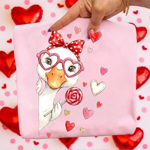 May include: Pink fabric featuring a cartoon goose wearing heart-shaped glasses and a red bow. The goose holds a lollipop and is surrounded by heart illustrations. The background includes red heart-shaped balloons and pink polka dots.