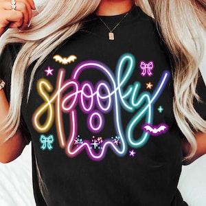 Op de afbeelding: Zwart T-shirt met een neon "Spooky" ontwerp. Het woord "Spooky" is geschreven in een speels lettertype met een spookomtrek en is omgeven door vleermuizen, sterren en strikken. Het ontwerp gebruikt gele, roze, paarse en turquoise kleuren.