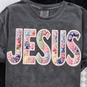 Könnte beinhalten: Dunkelgraues Comfort Colors T-Shirt mit dem Wort "JESUS" auf der Vorderseite. Die Buchstaben bestehen aus floralem Patchwork-Stoff mit weißem Perlenbesatz. Das Shirt hat einen Rundhalsausschnitt und kurze Ärmel.