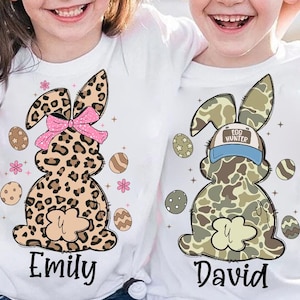 May include: Two white t-shirts with Easter bunny designs. One bunny is leopard print with a pink bow and the name "Emily". The other bunny is camouflage with a blue baseball cap that says "Egg Hunter" and the name "David".