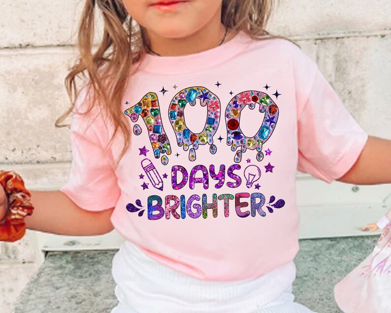 Op de afbeelding: Roze T-shirt met een kleurrijke afbeelding met de tekst "100 dagen stralender". De afbeelding bevat een getal 100 gemaakt van kleurrijke juwelen en een potlood en een gloeilamp.