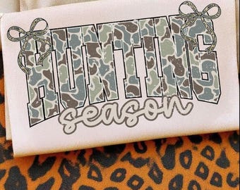 May include: A white t-shirt with the words "HUNTING SEASON" in a camouflage pattern. The letters are outlined in black and have a bow on each side. The shirt is on a background with a leopard print.