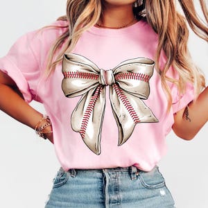 May include: A pink t-shirt with a white baseball-stitched bow on the front.