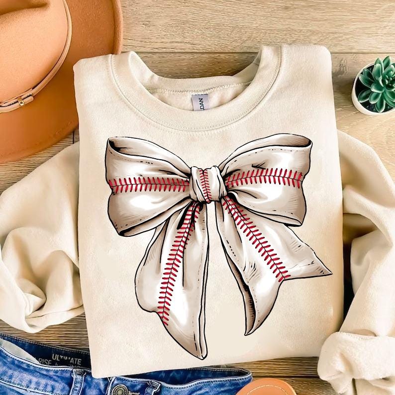 May include: A cream-colored sweatshirt with a large white bow with red stitching, resembling a baseball. The bow is centered on the front of the sweatshirt.