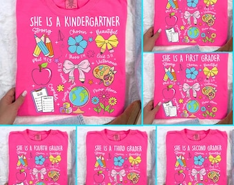 May include: A collection of pink t-shirts with colorful graphics and text. One shirt says "She is a Kindergartner" with school-themed illustrations. Other shirts are labeled "Preschooler", "First Grader", "Second Grader", "Third Grader", and "Fourth Grader".