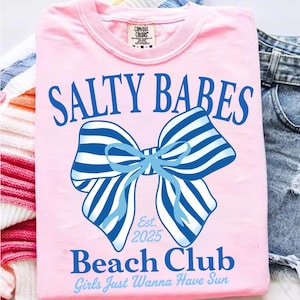 Może przedstawiać: R&oacute;żowa koszulka z niebieskim napisem "SALTY BABES" nad niebiesko-białą, pasiastą kokardą. Pod kokardą znajduje się napis "Beach Club" i "Girls Just Wanna Have Sun". Koszulka ma okrągły dekolt.