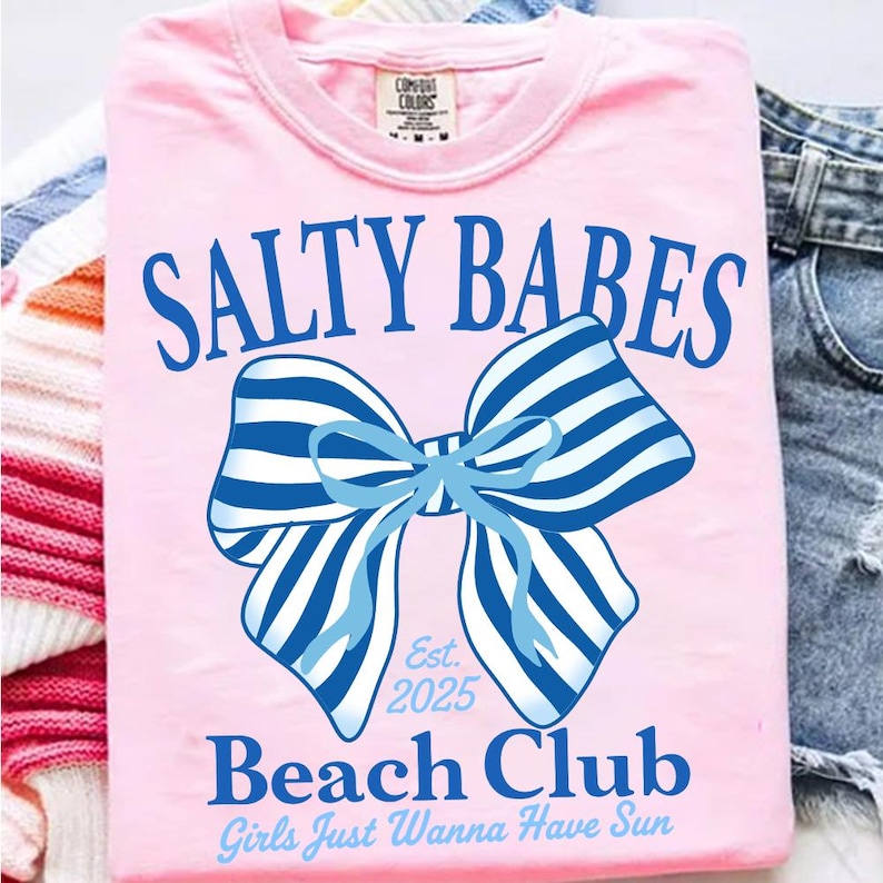 Może przedstawiać: R&oacute;żowa koszulka z niebieskim napisem "SALTY BABES" nad niebiesko-białą, pasiastą kokardą. Pod kokardą znajduje się napis "Beach Club" i "Girls Just Wanna Have Sun". Koszulka ma okrągły dekolt.