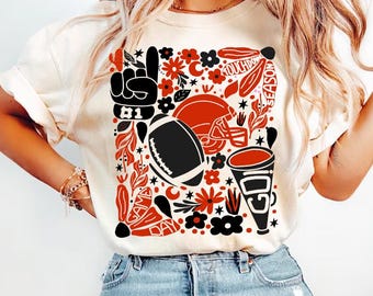 May include: Cream-colored t-shirt featuring a football-themed graphic. The design includes a football, helmet, and cheerleading megaphone in black and red, with floral and leaf accents. The words "Touchdown Season" are also visible.