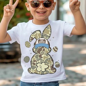 May include: White t-shirt featuring a cartoon camouflage bunny wearing a blue and white baseball cap that says "Egg Hunter". The bunny is surrounded by colourful Easter eggs.