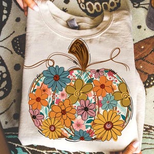May include: A beige sweatshirt featuring a floral pumpkin design. The pumpkin is outlined in brown with a brown stem and filled with colorful flowers in shades of blue, orange, yellow, and pink. The sweatshirt is folded.