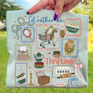 Puede incluir: Camiseta azul claro con el texto "I'd rather be thrifting" e ilustraciones variadas de artículos vintage, incluyendo un carrito de compras, un búho y tazas de té. El diseño está enmarcado con bordes decorativos y detalles florales.