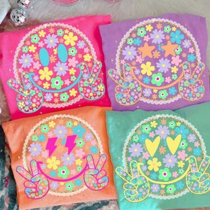 Könnte beinhalten: Vier farbenfrohe T-Shirts mit Smiley-Gesichtern. Jedes Shirt hat ein anderes Design: pink mit Blumen, lila mit Sternen, orange mit Blitzen und grün mit Herzen. Die Smiley-Gesichter haben Peace-Zeichen als Hände.