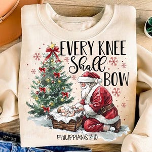 May include: A cream-colored sweatshirt with a Christmas-themed design. Santa Claus kneels beside a manger with a baby Jesus inside. A Christmas tree with red bows and ornaments is in the background. The text "Every Knee Shall Bow" is above the image and "Philippians 2:10" is below.