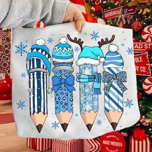 May include: A white pillow featuring four blue pencils with winter-themed hats, scarves, and bows. Each pencil has a different design, with snowflakes scattered around. The pencils are in a vertical arrangement, with a Christmas tree in the background.