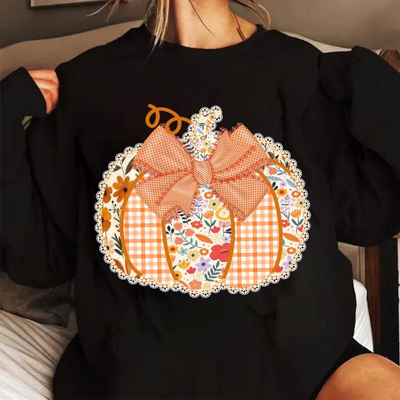 K&ouml;nnte beinhalten: Schwarzes Sweatshirt mit einem K&uuml;rbis-Design. Der K&uuml;rbis hat eine karierte Mitte, florale Seiten und eine gepunktete Schleife. Das Design ist mit einer wei&szlig;en Spitzentrimmung umrandet, ideal f&uuml;r Herbst.