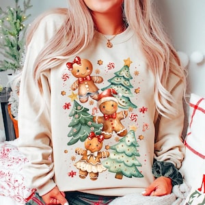 Könnte beinhalten: Ein beiger Pullover mit einem weihnachtlichen Design mit Lebkuchenmännern und Weihnachtsbäumen. Die Lebkuchenmänner tragen rote Schleifen und Weihnachtsmützen. Die Bäume sind mit Zuckerstangen und Sternen verziert.