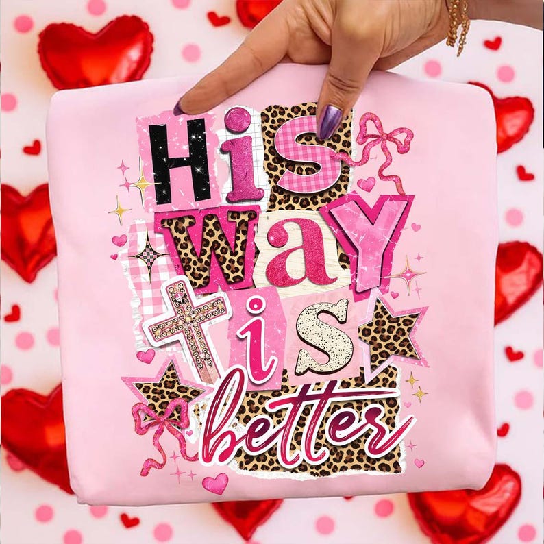 May include: A light pink t-shirt with the text "His Way is Better" in a collage style. The design includes a cross, stars, and leopard print accents. The background features red heart-shaped balloons.