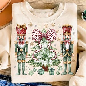 Puede incluir: Una sudadera beige con un diseño navideño con dos cascanueces flanqueando un árbol de Navidad verde con un lazo rojo y blanco de lunares. El árbol está decorado con bastones de caramelo y piruletas.
