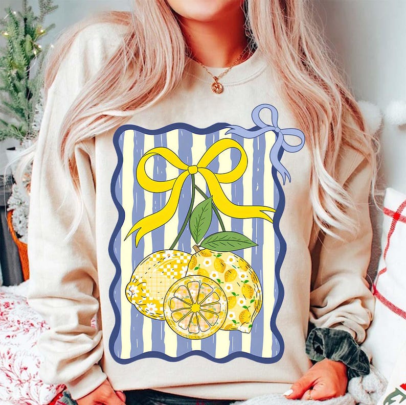 Pu&ograve; includere: Una felpa color crema con una grafica di due limoni con un fiocco giallo su uno sfondo a righe blu e bianco.