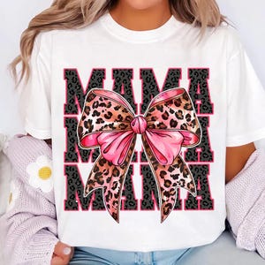 Pode incluir: Camiseta branca com um la&ccedil;o de estampa de leopardo rosa e marrom e a palavra "MAMA" repetida tr&ecirc;s vezes em texto preto.