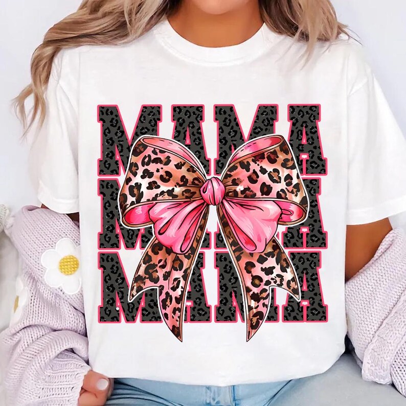 Pode incluir: Camiseta branca com um la&ccedil;o de estampa de leopardo rosa e marrom e a palavra "MAMA" repetida tr&ecirc;s vezes em texto preto.