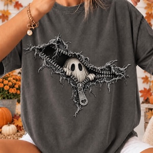 May include: A charcoal gray t-shirt featuring a graphic print of a white ghost peeking out from behind a realistic, open metal zipper design. The zipper pull is shaped like a small heart, and the fabric around the zipper appears frayed.