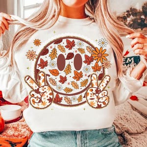 May include: White sweatshirt featuring a large smiley face design with fall leaves, daisies, and peace signs. The smiley face has brown eyes and a brown outline. The peace signs and the smiley face are decorated with fall leaves and flowers.