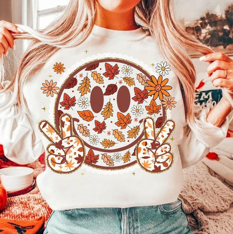 May include: White sweatshirt featuring a large smiley face design with fall leaves, daisies, and peace signs. The smiley face has brown eyes and a brown outline. The peace signs and the smiley face are decorated with fall leaves and flowers.