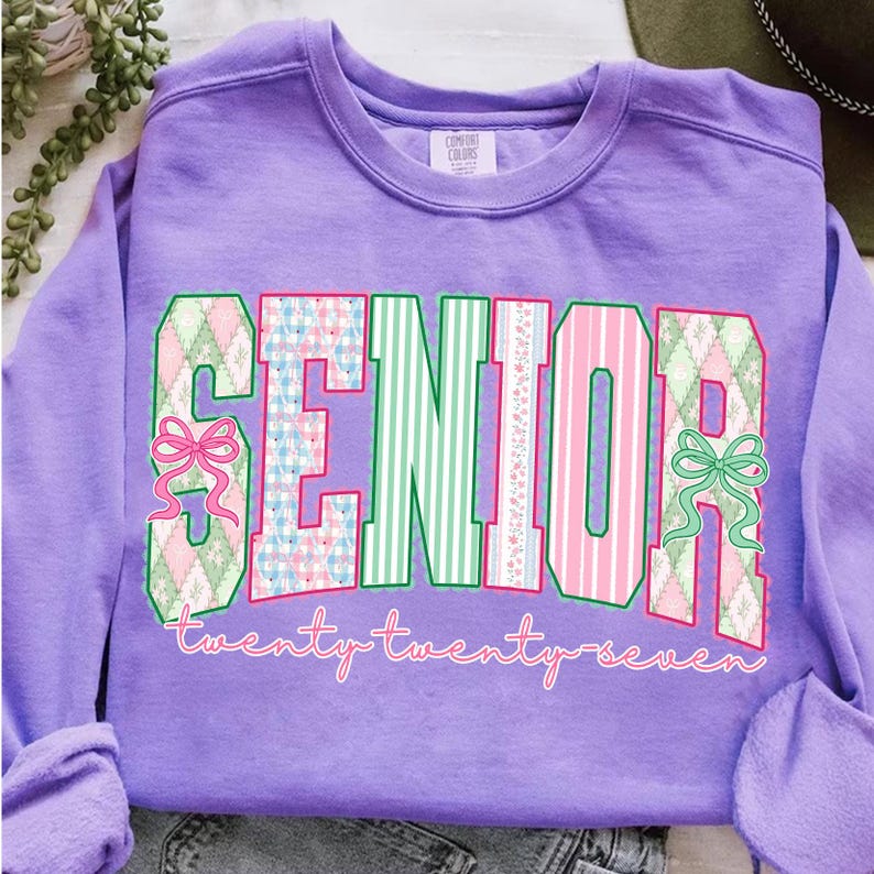 May include: A lilac-colored sweatshirt with the word "SENIOR" in large, colorful letters. The letters are decorated with patterns of green, pink, and blue, and bows. The text "twenty twenty seven" is written in cursive below the word "SENIOR".