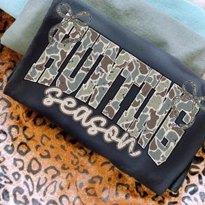 May include: A dark gray t-shirt with the words "HUNTING SEASON" in a camouflage pattern. The design includes decorative bows and is set against a backdrop of folded shirts and a leopard print.
