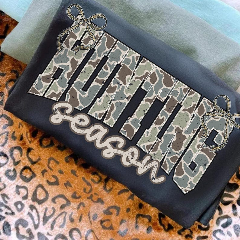 May include: A dark gray t-shirt with the words "HUNTING SEASON" in a camouflage pattern. The design includes decorative bows and is set against a backdrop of folded shirts and a leopard print.