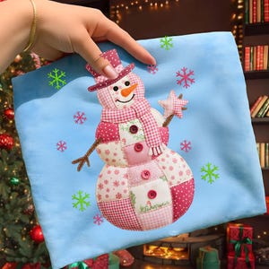 May include: A light blue cushion with a patchwork snowman design. The snowman has a carrot nose, a pink and white patterned hat and scarf, and holds a star. Decorative snowflakes surround the snowman. A festive holiday decoration.