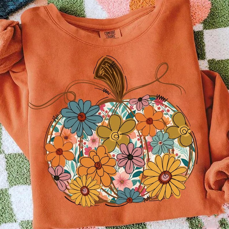 May include: An orange sweatshirt with a floral pumpkin design. The pumpkin is filled with colourful flowers in shades of orange, yellow, blue, and pink. The sweatshirt has a crew neck and long sleeves, ideal for autumn.