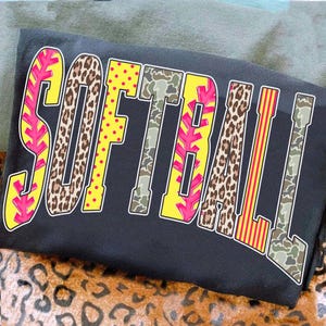 May include: Black sweatshirt featuring the word "SOFTBALL" in large, patterned letters. The letters incorporate leopard print, camouflage, and yellow with pink details. The garment is folded on a leopard print surface, suggesting a casual style.
