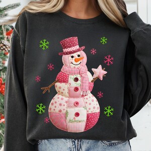 May include: Dark grey sweatshirt with a patchwork snowman design. The snowman has a burgundy top hat, scarf, and buttons, with a carrot nose and a star-shaped wand. Pink and green snowflakes surround the snowman.
