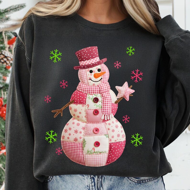 May include: Dark grey sweatshirt with a patchwork snowman design. The snowman has a burgundy top hat, scarf, and buttons, with a carrot nose and a star-shaped wand. Pink and green snowflakes surround the snowman.