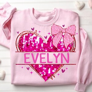 May include: Pink sweatshirt with a sequined heart and a pink bow. The heart has the name "EVELYN" written on it.