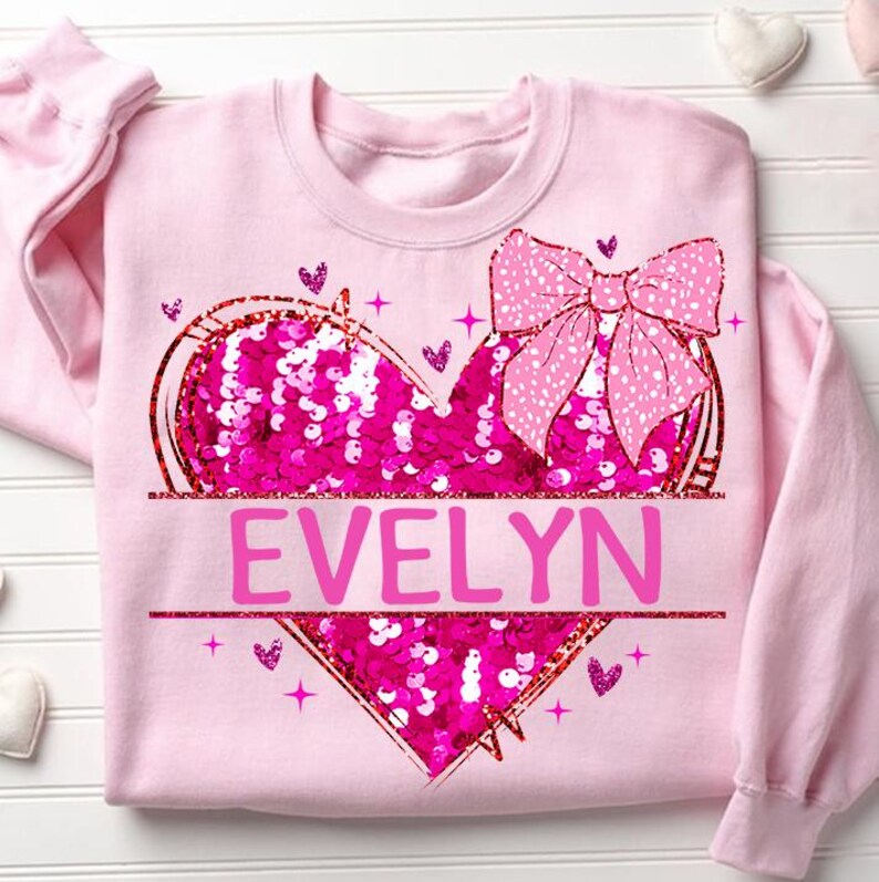 May include: Pink sweatshirt with a sequined heart and a pink bow. The heart has the name "EVELYN" written on it.