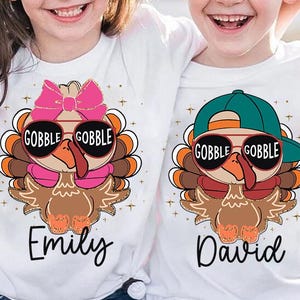 May include: Two white t-shirts with a Thanksgiving turkey design. The turkey is wearing sunglasses and a pink bow or a green baseball cap. The shirts are personalized with the names "Emily" and "David".