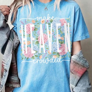 May include: A light blue t-shirt with the words "make HEAVEN crowded" in white. The word "HEAVEN" is in large capital letters, with a floral design of pink, blue, and peach flowers behind it.