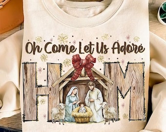 May include: A beige sweatshirt with a Christmas nativity scene design. The design features a wooden manger with a red bow, a baby Jesus, Mary, Joseph, and two sheep. The text "Oh Come Let Us Adore Him" is written above the scene.