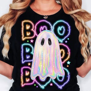Pu&ograve; includere: T-shirt nera con un design neon "BOO" colorato e una grafica di fantasmi pastello. Il design include scritte al neon e stelle, perfetto per Halloween o la stagione spettrale.