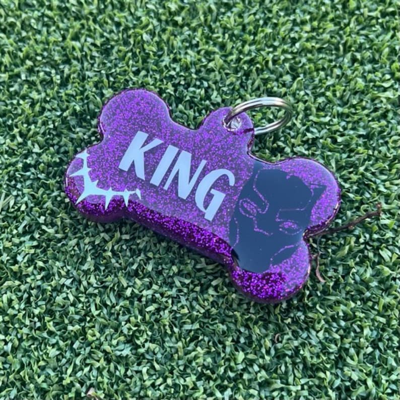 May include: A bone-shaped pet tag with a purple glitter finish, featuring the word KING in bold white letters and a black graphic of a panther mask on the right side, complete with a silver metal split ring for attachment to a collar.