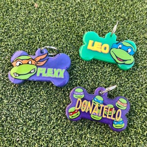 May include: Three purple and green bone-shaped dog tags with Teenage Mutant Ninja Turtles characters and names. The tags read "Flexx", "Donatello", and "Leo".