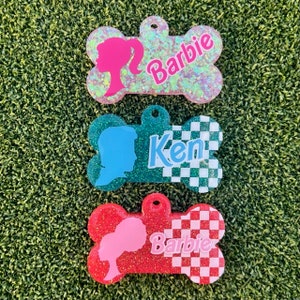 May include: Three bone-shaped dog tags with glitter and checkered patterns. The top tag is pink with a silhouette of a woman and the text "Barbie". The middle tag is teal with a silhouette of a man and the text "Ken". The bottom tag is red with a silhouette of a woman and the text "Barbie".