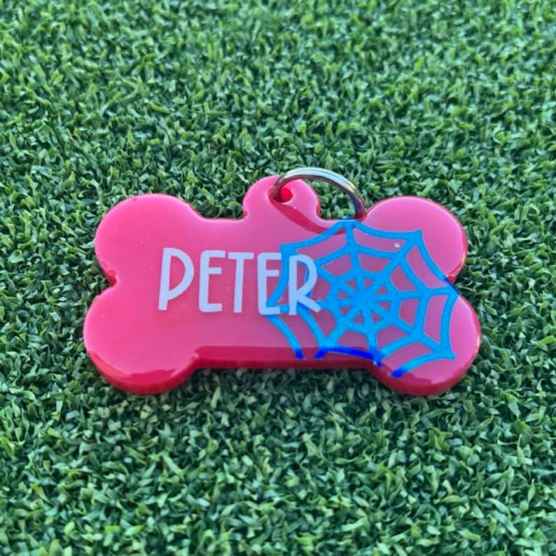 May include: A bright pink bone-shaped pet tag featuring the name PETER in white capital letters and a blue spiderweb graphic on the right side. The tag includes a silver metal jump ring attached to the top loop for secure collar attachment.