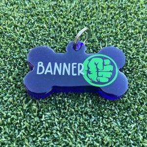 May include: A dark blue, bone-shaped pet identification tag featuring the white text BANNER and a green circular graphic of a clenched fist. The tag includes a silver metal split ring attached to the top loop for secure collar attachment.