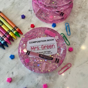 May include: Pink glitter resin coaster with a white label that says "Composition Book Mrs. Green 100 Sheets 200 Pages 24.7 x 19.0 cm Crayola". The coaster also has a pink eraser, a white push pin, a pink crayon, and a pink paper clip.