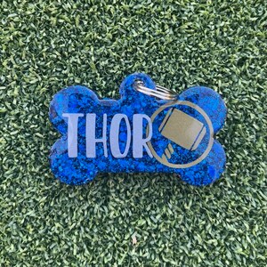 May include: A bone-shaped pet ID tag featuring a deep blue glitter finish. The name THOR is printed in large, light gray capital letters across the center. To the right of the name is a circular gold icon depicting a hammer. A metal ring is attached.