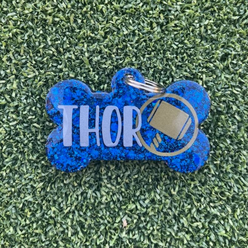 May include: A bone-shaped pet ID tag featuring a deep blue glitter finish. The name THOR is printed in large, light gray capital letters across the center. To the right of the name is a circular gold icon depicting a hammer. A metal ring is attached.